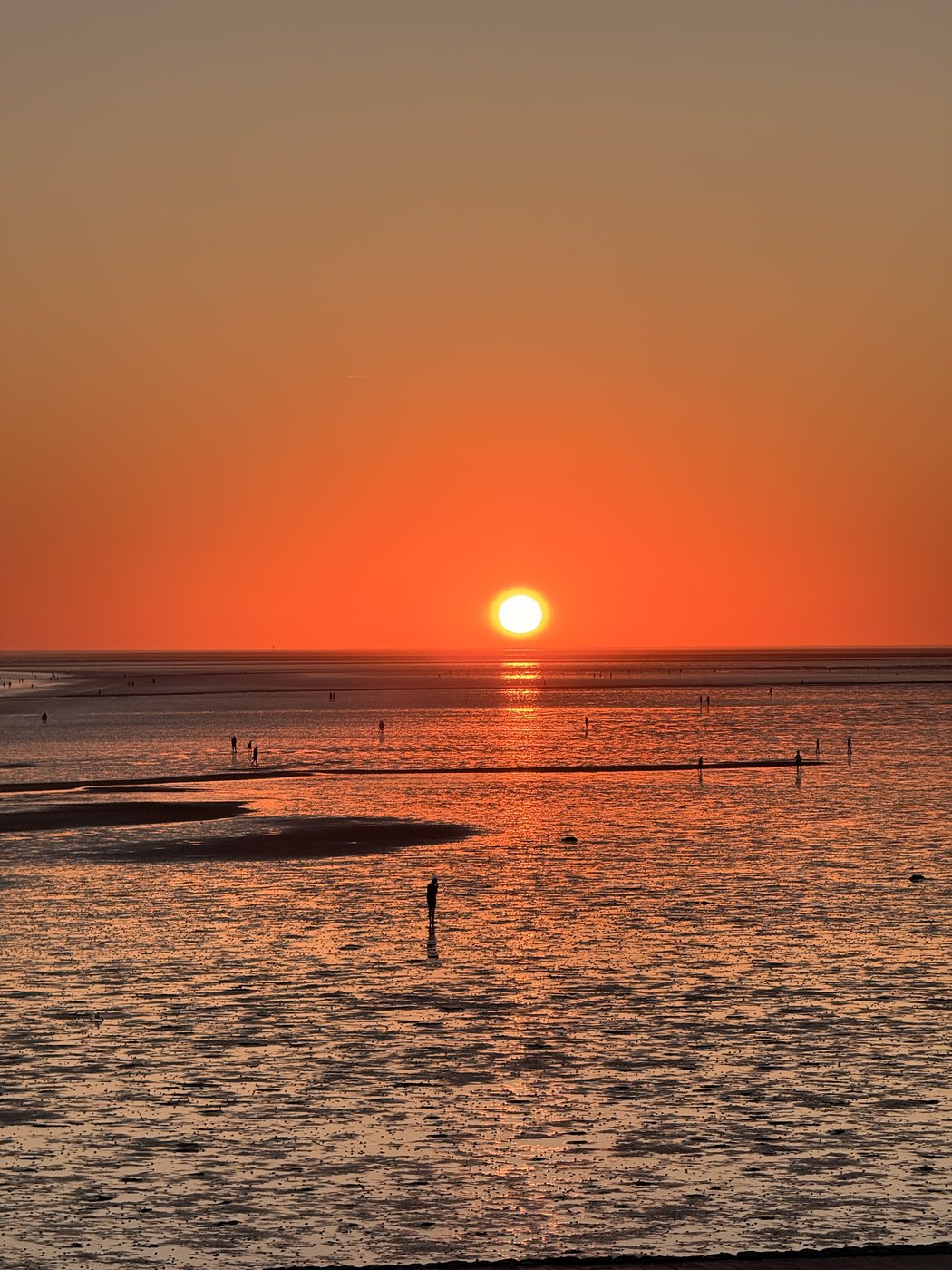 Die Sonne geht über dem Wattenmeer unter, kleine Gestalten laufen im flachen Wasser